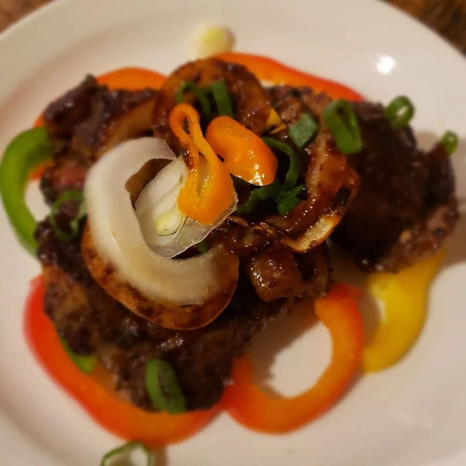 Jamaican dish with dark meat, colorful bell peppers, onions, and green garnishes on a white plate.