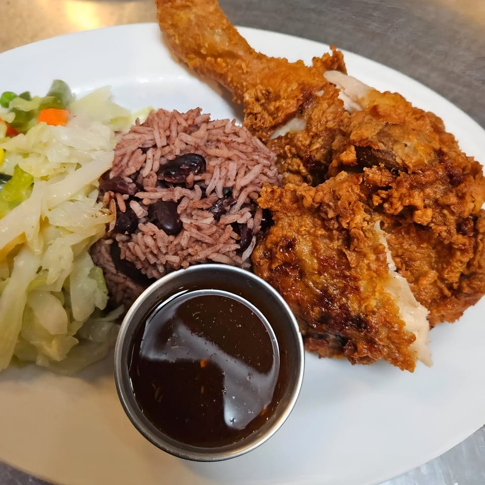 Crispy fried chicken, rice and peas, steamed cabbage, and a side of sauce on a white plate.