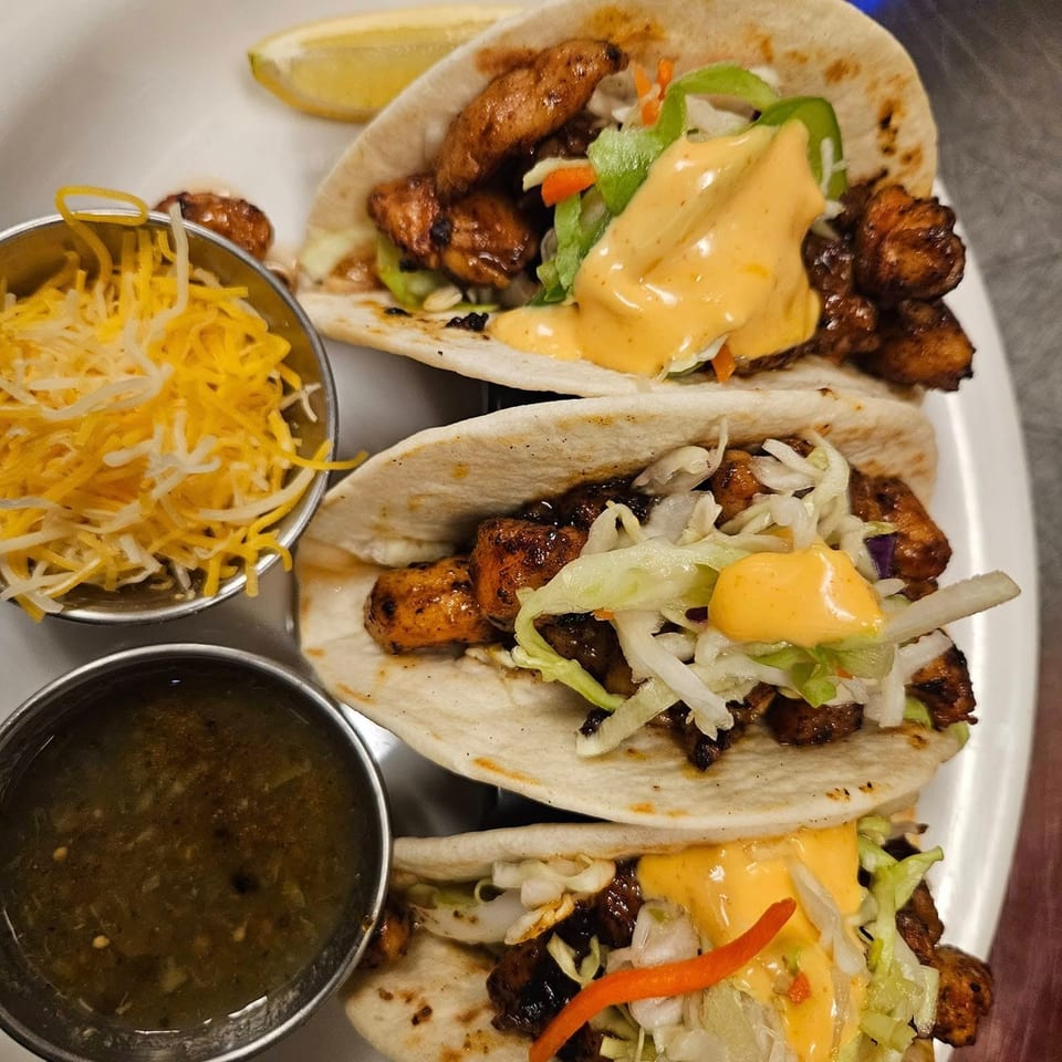 Three jerk chicken tacos with shredded cabbage, sauce, and sides of cheese and salsa.