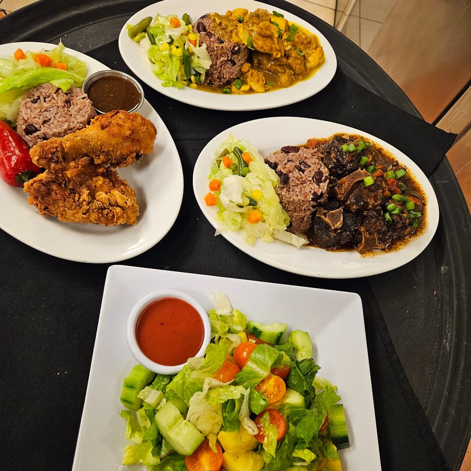 Four plates of Jamaican food: fried chicken, curry chicken, oxtail, rice and peas, and salads.