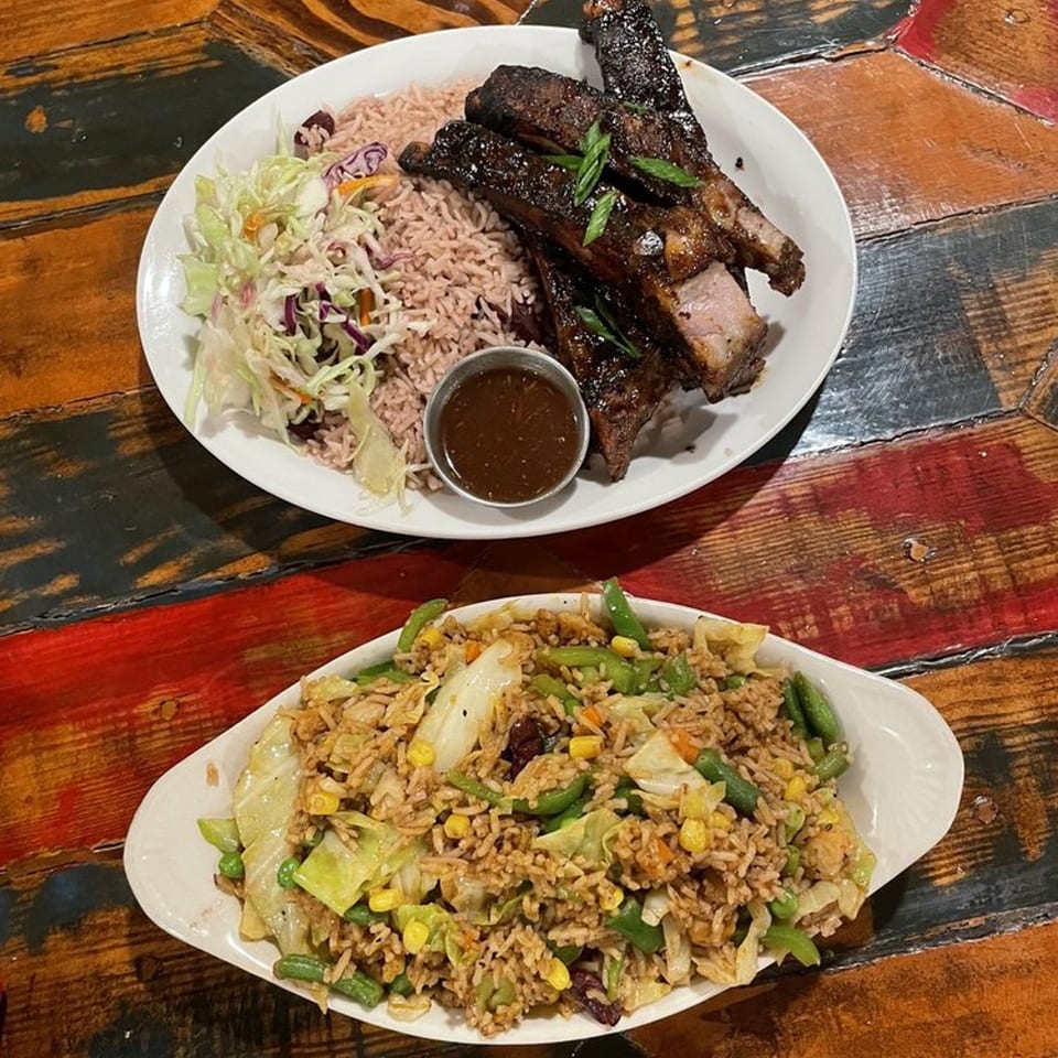 Two plates of Jamaican food: jerk ribs with rice and coleslaw, and vegetable fried rice.