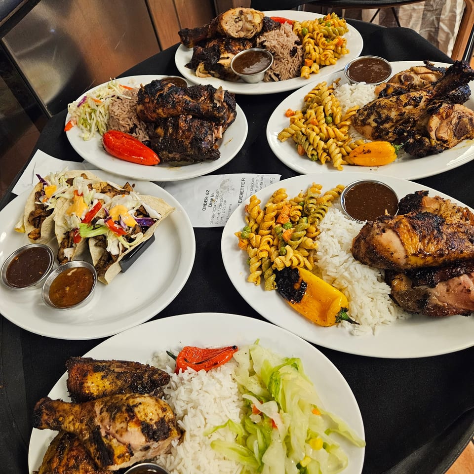 Multiple plates of Jamaican jerk chicken, rice, pasta salad, and tacos on a black tablecloth.